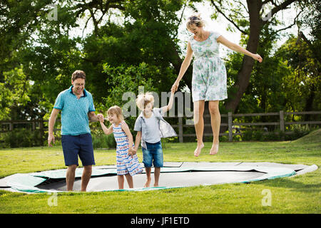 Homme, Femme, garçon et fille tenant les mains, sauter sur un trampoline installé dans la pelouse dans un jardin. Banque D'Images