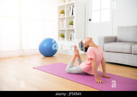 Little girl doing yoga exercice sur tapis de sol en bois avec les jambes et le corps en flexion studio de remise en forme près de canapé avec de grandes fenêtres sur l'arrière-plan dans l'livin Banque D'Images