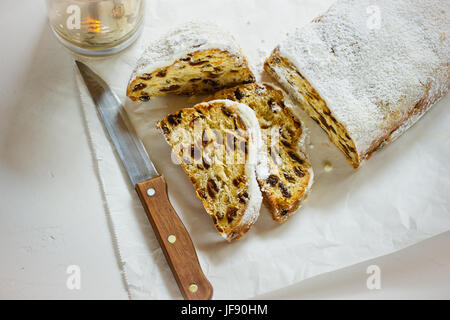 Stollen de Noël allemand avec des tranches coupées sur le papier parchemin, fond en béton blanc, couteau, bougie allumée, propre, de style minimaliste, vue du dessus Banque D'Images