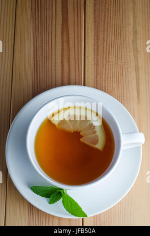 Coup de frais généraux de thé au citron dans une tasse et soucoupe blanche avec une demi-tranche de citron et de feuilles de menthe sur une table en bois recouvert. Banque D'Images