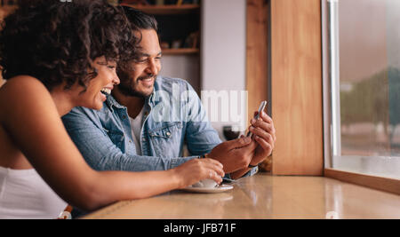 Smiling young couple sitting at coffee shop looking at mobile phone. Jeune homme et femme au café à l'aide de smart phone. Banque D'Images