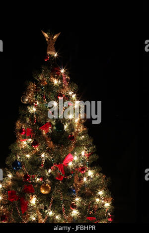 Peu mousseux rouge arbre de Noël avec des arcs, des décorations colorées et éclairées angel sur le haut,isolé sur un fond noir avec l'exemplaire de l'espace. Banque D'Images