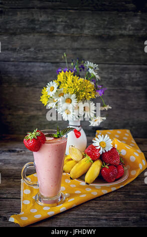 Smoothie fraise-banane avec du blanc à pois colorés avec bol de fraises sur une serviette jaune et d'un vase de fleurs sauvages . Sol en bois backgroun Banque D'Images