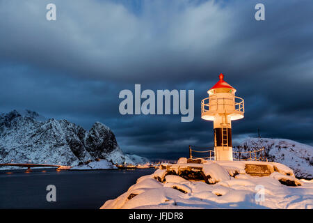 Un phare dans la neige dans la nuit de l'Arctique avec le village de Reine dans l'arrière-plan le Nordland Lofoten, Norvège Europe Banque D'Images