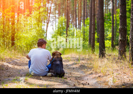 Homme assis avec un chien sur la route de terre dans une forêt de pins en été retour à l'appareil photo Banque D'Images