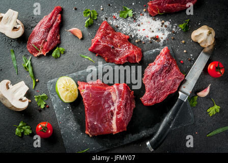 La viande de boeuf cru frais filet mignon sur une planche à découper ardoise sur noir table de pierre. Avec les épices (sel, poivre), de l'ail et fines herbes - persil et tarrag Banque D'Images