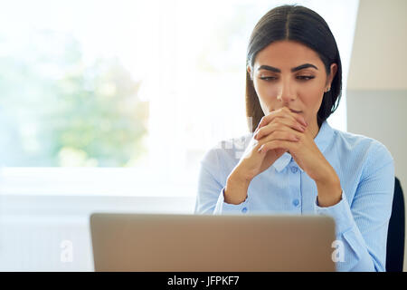 Young businesswoman sérieuse avec une expression inquiète en train de lire l'information sur son ordinateur portable avec les mains jointes contre une fenêtre haute avec spa de copie Banque D'Images