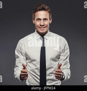 Smiling young businessman portant une chemise et cravate de donner un pouce levé debout seul dans un studio sur un arrière-plan gris Banque D'Images