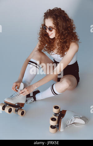 Portrait de jeune fille portant des lunettes de hipster en patins à assis isolé sur gray Banque D'Images