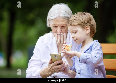 Jeune garçon et de sa grand-mère à l'aide de smartphone pendant que sitting on bench in park Banque D'Images