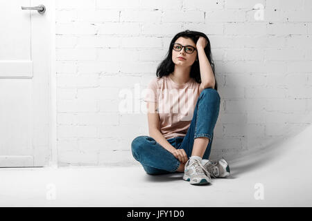 Portrait de smart belle brunette girl à lunettes avec le maquillage naturel blanc assis près de mur de briques. Banque D'Images