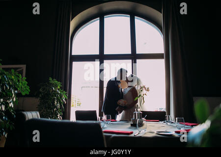 Beau, romantique newlyweds kissing près de fenêtre lumineuse dans la salle de mariage avant la célébration Banque D'Images