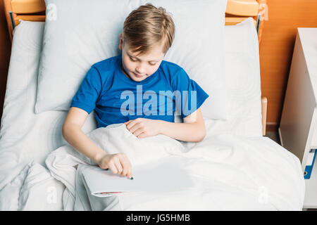 Smiling little boy drawing while lying in hospital bed Banque D'Images