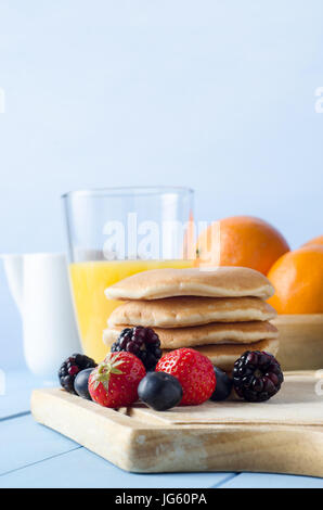 Une table de cuisine avec des crêpes empilées sur planche en bois entourée de fruits d'été, un verre de jus d'orange, bol de lait et d'oranges Banque D'Images