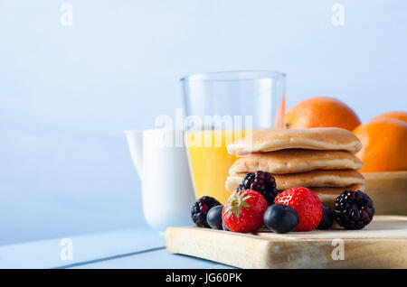 Scène d'été avec table de petit déjeuner des fruits et une pile de crêpes sur une vieille planche en bois, un bol d'oranges, jus d'orange dans une gla Banque D'Images