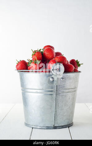 Vue latérale d'une vieille, dénoyautées et rayé aluminium metal seau rempli à craquer avec des fraises rouges récoltés sur un livre blanc de planches en bois peint Banque D'Images