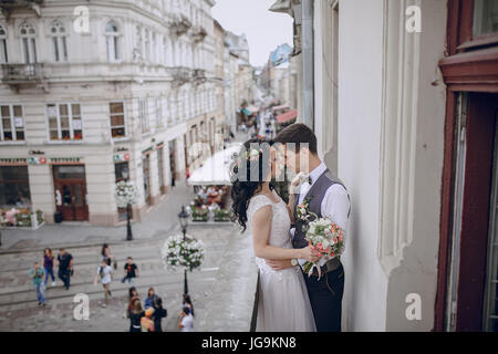 Mariée en robe blanche debout sur un balcon donnant sur la ville et biket tient une fleur à côté de son groom standing en chemise blanche Banque D'Images