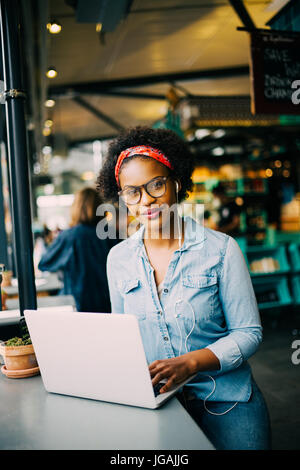 Souriante jeune femme africaine assis seul au comptoir dans un bistrot de travailler sur un ordinateur portable et d'écouter de la musique sur l'earphones Banque D'Images