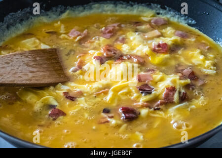 Oeufs brouillés au jambon et bacon par cuisson dans un poêlon noir, mélangé avec une louche en bois Banque D'Images