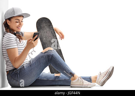 Adolescente avec une planche à roulettes à la recherche à un téléphone et appuyé contre un mur isolé sur fond blanc Banque D'Images