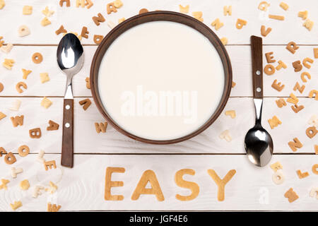 Vue rapprochée du lait dans un bol, deux cuillères, mot facile de céréales petit déjeuner lettres alphabet et flocons de maïs sur la table en bois Banque D'Images