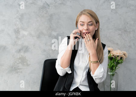 Teen Caucasian woman in casual vêtements est talking on mobile phone et montrant surprise tout en restant assis à la maison. Banque D'Images