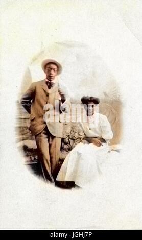 Couple afro-américains, un homme et une femme, se faisant passer pour un studio portrait contre une toile peinte, l'homme en costume et chapeau de paille et debout, la femme vêtue d'une robe blanche et assis avec les bras croisés sur ses genoux, à la fois avec les expressions du visage grave, 1905. Remarque : l'image a été colorisée numériquement à l'aide d'un processus moderne. Les couleurs peuvent ne pas être exacts à l'autre. Banque D'Images