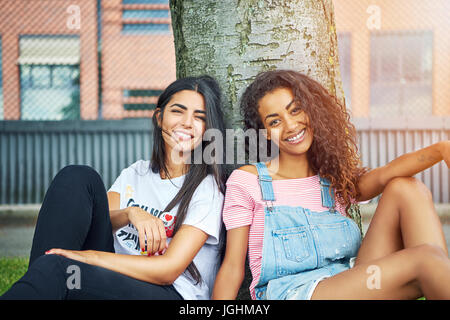 Deux jeunes amis attrayant tout en souriant, assis sur une pelouse, se penchant en arrière sur un tronc d'arbre sur une journée ensoleillée Banque D'Images