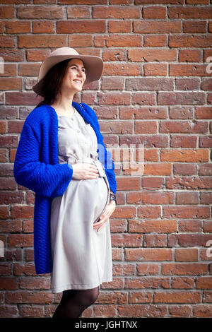 Une élégante femme enceinte dans une robe beige, un cardigan bleu et un chapeau se tient avec les mains sur son ventre dans le contexte d'un bâtiment en brique Banque D'Images