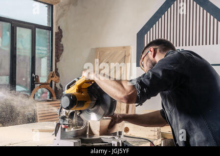 Un jeune menuisier construction moderne scies scie circulaire avec une planche en bois dans l'atelier, une sciure de bois voler sur les côtés, une vie arrière Banque D'Images