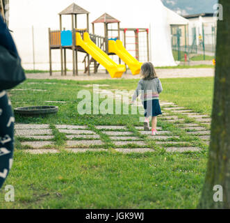 Petit bébé marche loin de sa mère et aller vers un château avec toboggan jouet dans un parc public, en cours d'exécution sur l'herbe (avec le dos face à nous) Banque D'Images