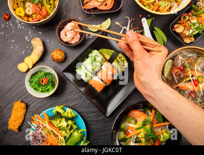 Man hand holding baguettes et d'échantillonnage des aliments asiatiques table avec différents types de plats chinois, nouilles, poulet, porc, boeuf, riz, soupe aigre, printemps rol Banque D'Images