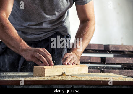 Close-up : un jeune male carpenter dans un t-shirt gris et de travail dans l'ensemble vole un bar en bois avec une fraiseuse à l'atelier Banque D'Images