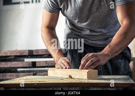 Close-up : un jeune homme carpenter dans un t-shirt gris et de travail dans l'ensemble vole un bar en bois avec une fraiseuse à l'atelier Banque D'Images