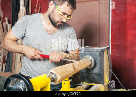 Un jeune homme brun un constructeur en vert lunettes de protection, un t-shirt gris et une barbe fait un produit en bois sur un tour à l'atelier Banque D'Images