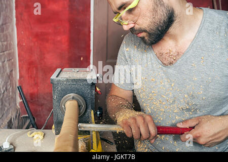 Une jeune brunette homme builder wearing green lunettes de protection, un t-shirt gris et avec une barbe de travailler sur une pièce en bois sur un tour dans le worksho Banque D'Images