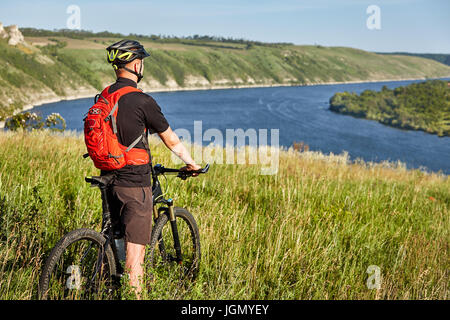 Vue arrière du jeune cycliste se tient avec vtt sur le pré vert au-dessus de Big River. Banque D'Images