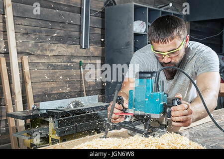 Un homme aux cheveux noirs et une barbe dans les lunettes de sécurité et un t-shirt gris est maintenant une fraiseuse et poignées d'une planche en bois dans l'atelier, à la Banque D'Images