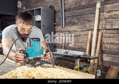 Un homme aux cheveux noirs et une barbe dans les lunettes de sécurité et un t-shirt gris est maintenant une fraiseuse et poignées d'une planche en bois dans l'atelier, à la Banque D'Images