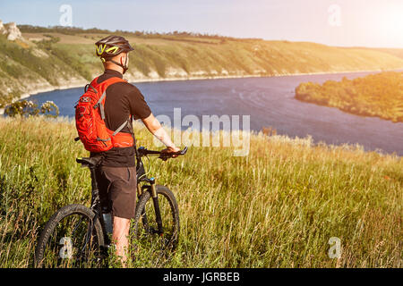 Vue arrière du jeune cycliste se tient avec vtt sur le pré vert au-dessus de Big River. Banque D'Images