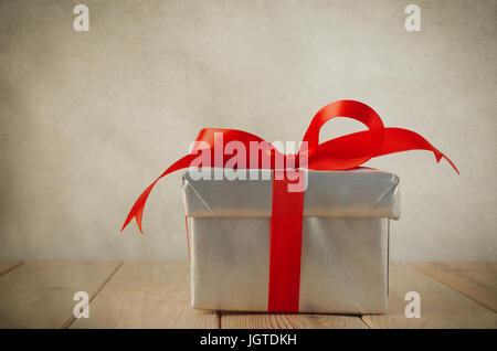 Un cadeau de Noël boîte avec couvercle fermé, enveloppé dans du papier alu et lié à un noeud avec un ruban de satin rouge. Placé sur une vieille table en bois avec Banque D'Images