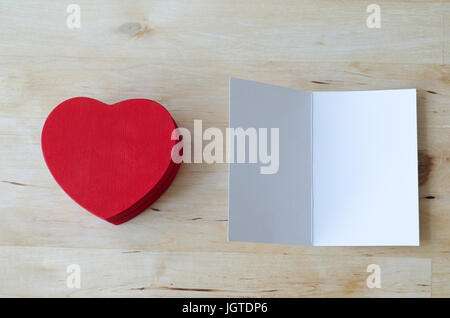 Passage tiré d'un rouge en forme de coeur en bois peint boîte cadeau et une carte de vœux, laissée en blanc pour un message. Les deux situé sur une table woodplank lumière Banque D'Images