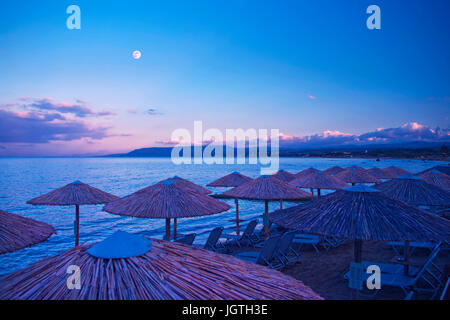 Des parasols et transats sur la plage la nuit avec les montagnes et la pleine lune en arrière-plan à Georgioupolis, Crète, Grèce Banque D'Images