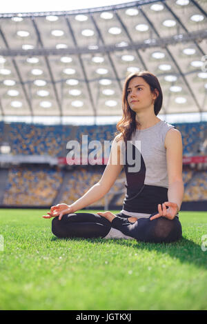 Jeune fille sportive heureux assis dans yoga pose dans le stade. Yoga woman meditating on Green grass contre le soleil Banque D'Images