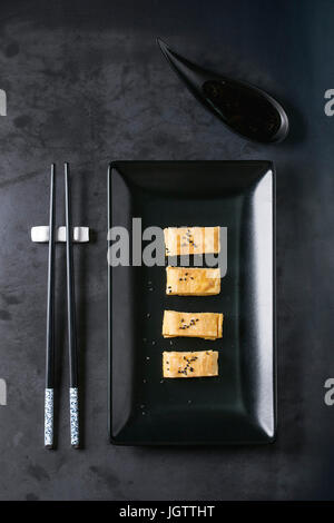 Omelette Tamagoyaki laminées japonais noir en tranches avec la sauce soja et les graines de seasame, servi dans carré noir plaque en céramique avec des baguettes plus dark metal Banque D'Images