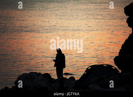 Silhouette de trekker sur rocky seashore près de camping tente sur nuit à coucher du soleil d'été Banque D'Images