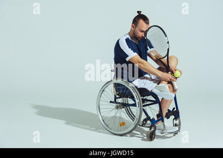 Joueur de tennis bouleversé assis en fauteuil roulant et holding tennis racquet ball et isolé sur gray Banque D'Images