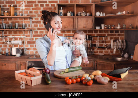 Jeune mère tenant son fils sur les armes et de parler sur smartphone pendant le dîner dans la cuisine de préparation Banque D'Images