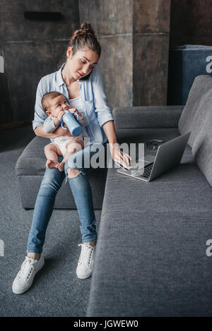 Jeune maman avec bébé garçon et de travail à distance en utilisant un ordinateur portable pendant une conversation sur smartphone Banque D'Images