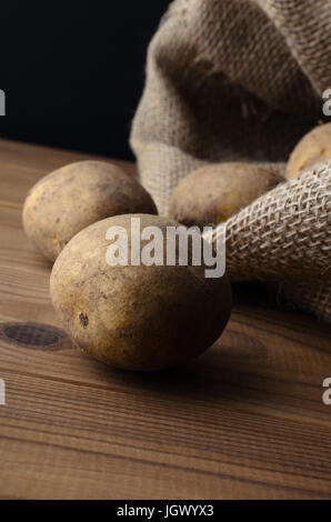 Matières, sans les laver, éplucher les pommes de terre, s'échappant de sac en toile de jute sur les planches de bois à table. Moody éclairage, fond noir. Banque D'Images